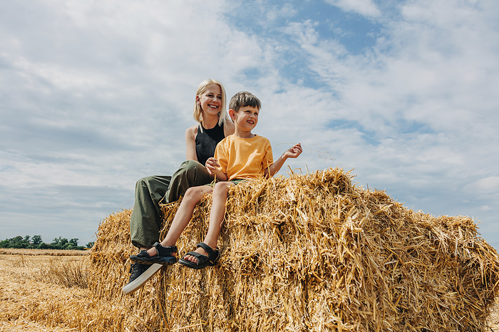 Smiling mother and son sitting on haystack in wheat field