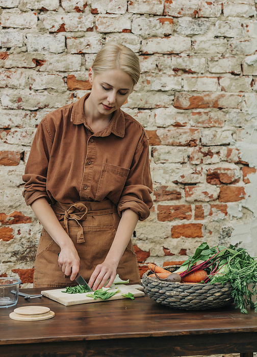 Blond woman cutting vegetables on table in front of brick wall