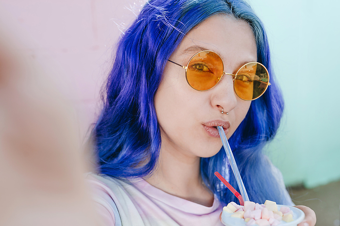 Hipster woman with dyed blue hair holding drink taking selfie at cafe
