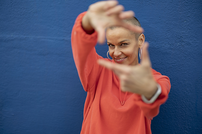 Smiling woman with shaved head showing finger frame in front of blue wall