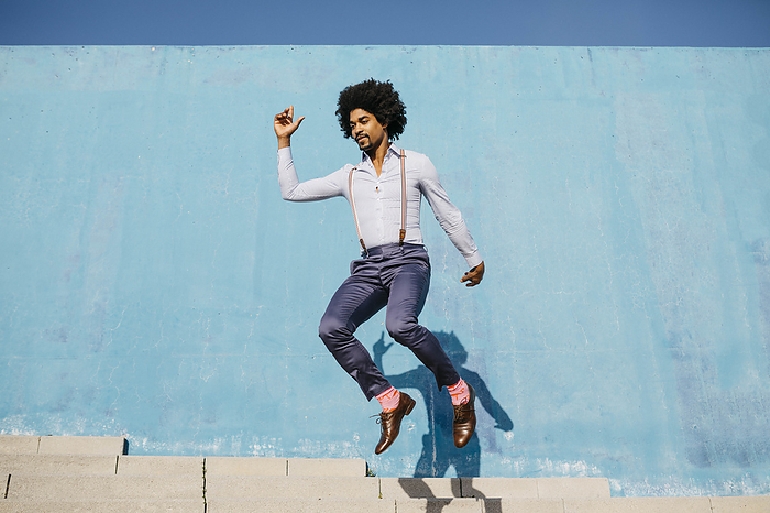 Stylish man jumping in the air in front of blue wall