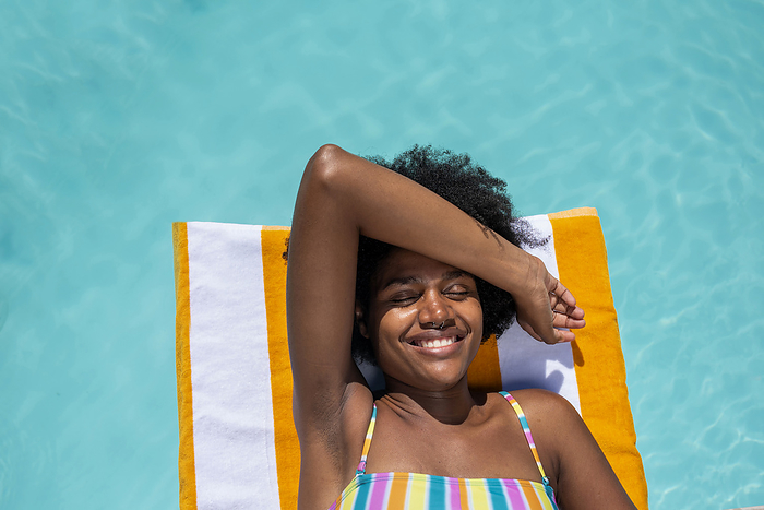Smiling young woman lying on stripped towel with eyes closed in pool
