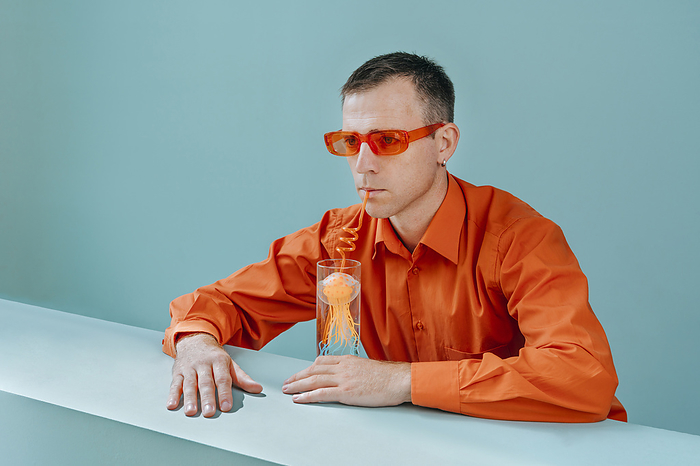 Man with orange sunglasses drinking water with jellyfish in glass against blue background