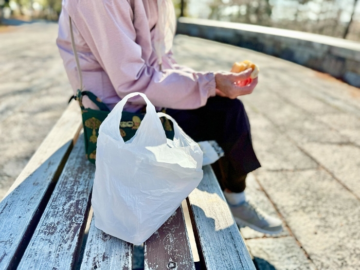 Elderly woman sitting on a bench with a plastic bag beside her eating a cream bun.
