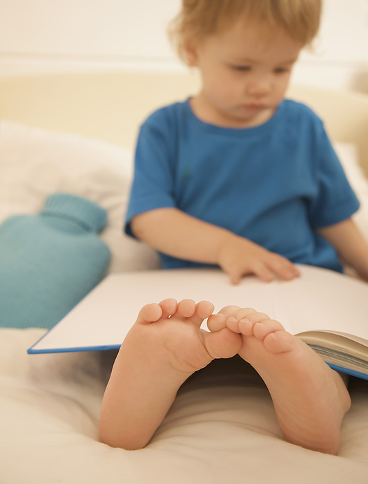 Young boy sitting in bed with an open book over is legs and feet, by Jutta Klee
