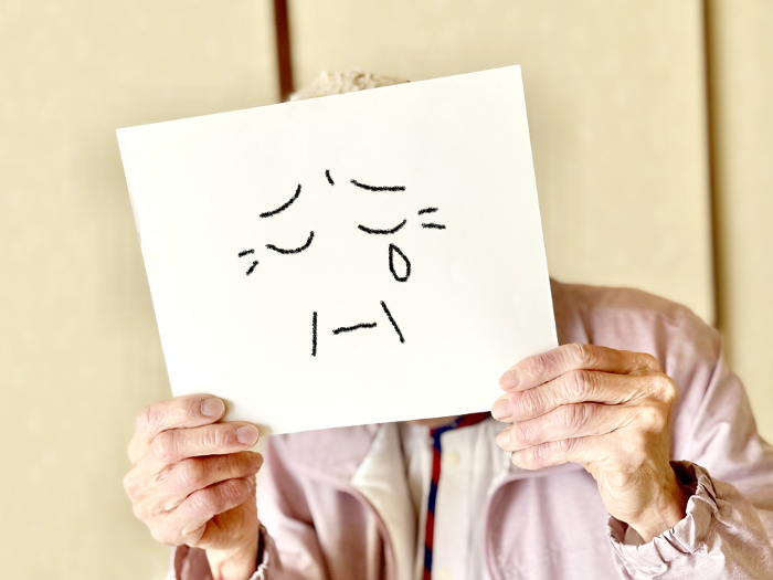 Elderly woman holding a piece of paper with illustrated face in front of her face with both hands, with tears in her eyes.