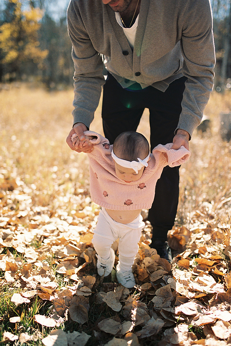 Dad helping baby girl wearing neutral clothing to walk in fall leaves, by Cavan Images / Samantha Joy Shantz Photography
