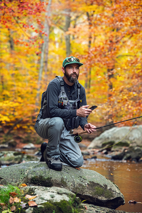 Man checking phone while fly fishing on a colorful fall day, by Cavan Images / Ben Wentzel