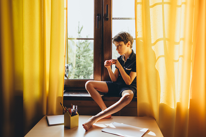 Quiet thoughtful boy sitting on a windowsill on a sunny day, by Cavan Images / Natalia Akulova
