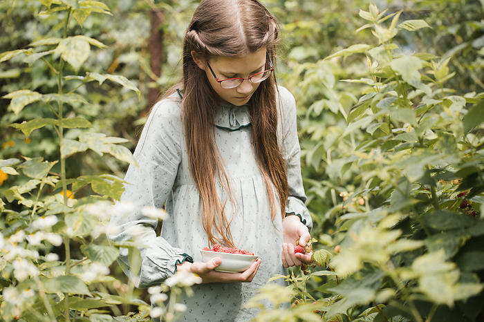 A teenage girl in glasses and a vintage dress picks raspberries, by Cavan Images / Iuliia Malivanchuk