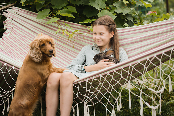 A teenage girl rests on a hammock with a rabbit and a dog, by Cavan Images / Iuliia Malivanchuk