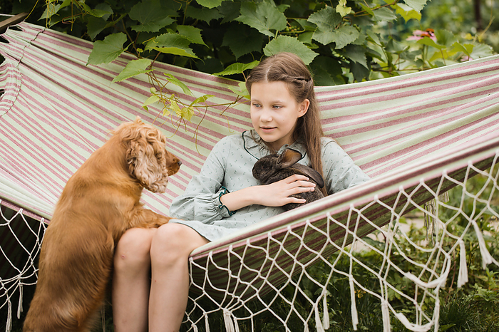 A girl rests on a hammock with a rabbit and a dog in the garden, by Cavan Images / Iuliia Malivanchuk