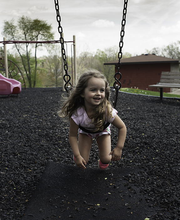 Little girl with long curls smiling on swing set, by Cavan Images / Joy Faith