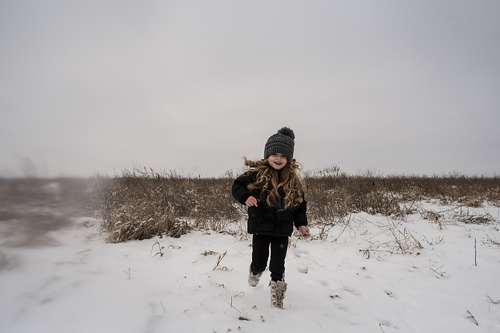 Little girl running through snowy field in boots and hat, by Cavan Images / Joy Faith