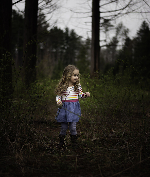 Beautiful little girl with blonde curls standing in pine forest, by Cavan Images / Joy Faith