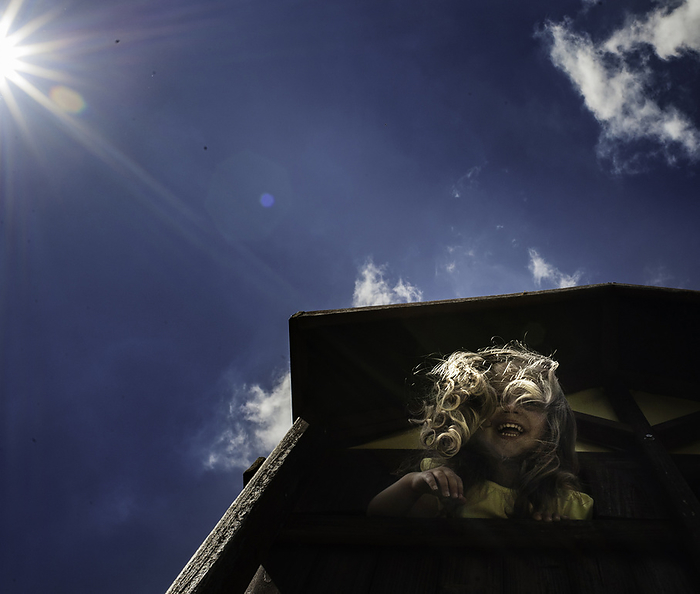 Happy little girl peeking over play house in summer, by Cavan Images / Joy Faith