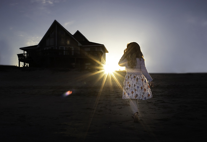 Little girl walking in sand towards house on beach at sunset, by Cavan Images / Joy Faith