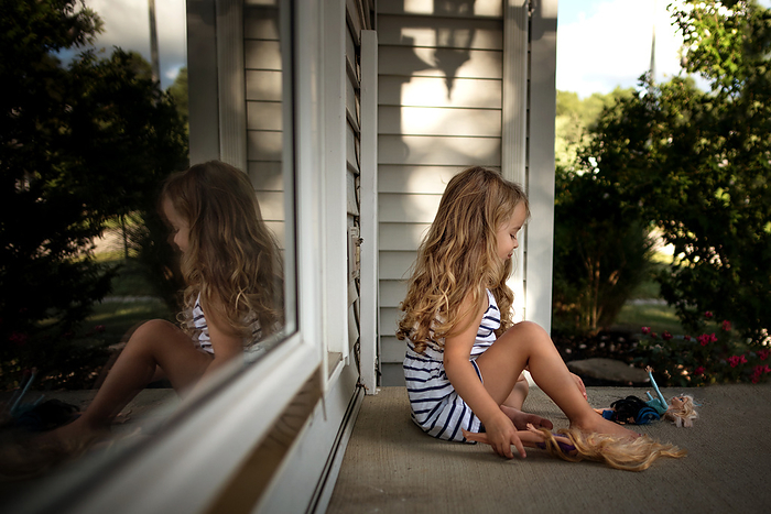 Little girl with long curls playing with dolls  on front porch, by Cavan Images / Joy Faith