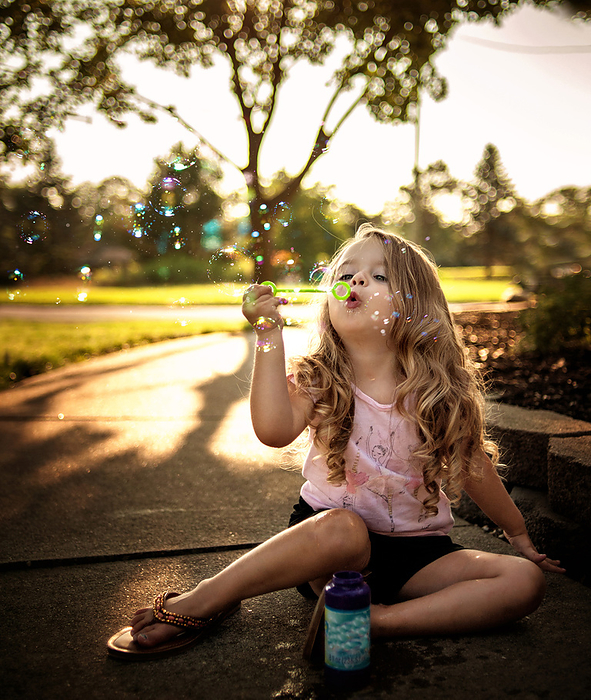 Toddler with long curls blowing bubbles outside, by Cavan Images / Joy Faith