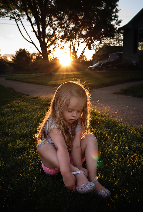 Cute little girl putting on shoes in yard at sunset, by Cavan Images / Joy Faith