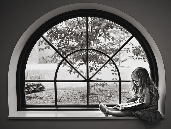 Little girl with long curls reading book in big window, by Cavan Images / Joy Faith