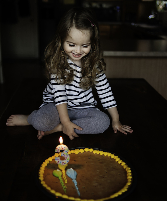 Happy little girl looking at birthday cake in kitchen, by Cavan Images / Joy Faith