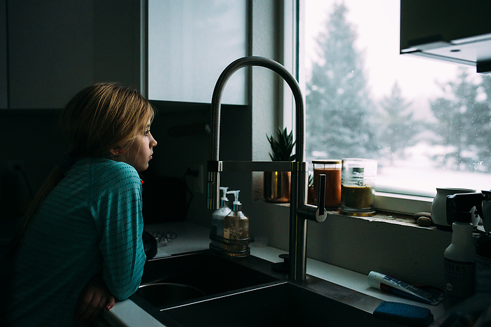 Preteen girl leans over kitchen sink and looks out window at sno, by Cavan Images / Kimberli Fredericks