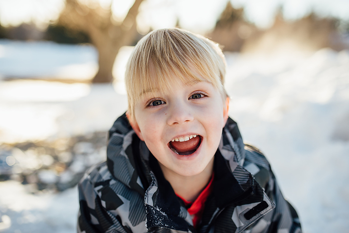 Young boy smiles mischeviously at camera outdoors with snow and, by Cavan Images / Kimberli Fredericks