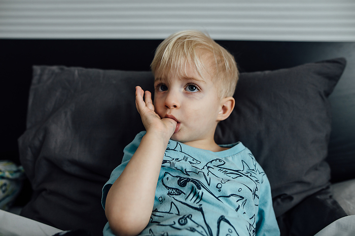 Close up of little boy sucking his thumb while sitting on bed at, by Cavan Images / Kimberli Fredericks