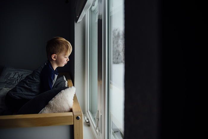 Small boy sits on bed and looks out window at snow below with dr, by Cavan Images / Kimberli Fredericks