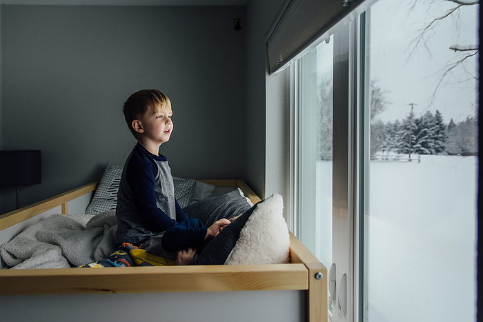 Bright side view of little boy sitting on bed looking out window, by Cavan Images / Kimberli Fredericks