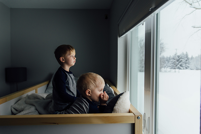 Bright side view of small brothers sitting on bed looking out wi, by Cavan Images / Kimberli Fredericks