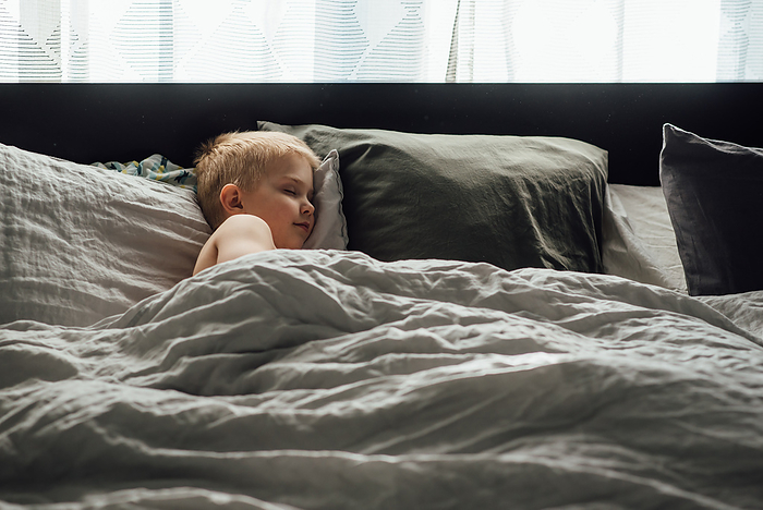 Wide view of small boy peacefully taking a nap on big bed with n, by Cavan Images / Kimberli Fredericks