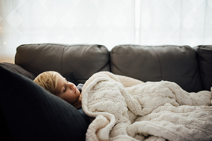 Side view of little boy napping on gray couch with white blanket, by Cavan Images / Kimberli Fredericks
