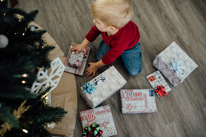 Little boy sits on ground surrounded by presents next to Christm, by Cavan Images / Kimberli Fredericks