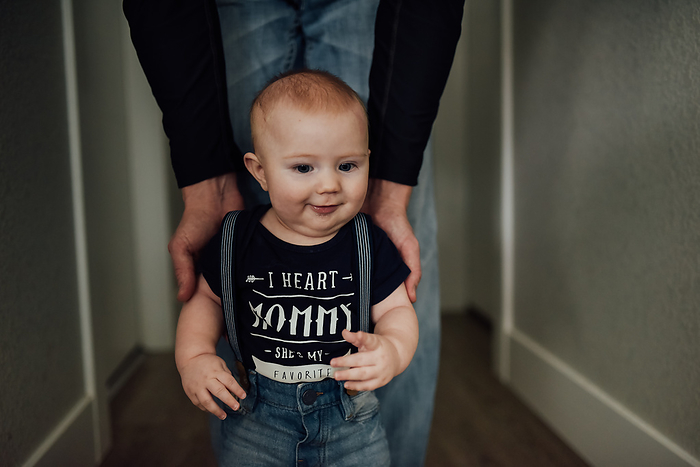 Dad helps baby boy stand in hallway at home., by Cavan Images / Kimberli Fredericks