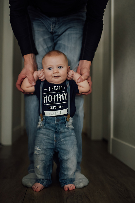 Baby boy wearing I heart mommy shirt stands in hallway while dad, by Cavan Images / Kimberli Fredericks