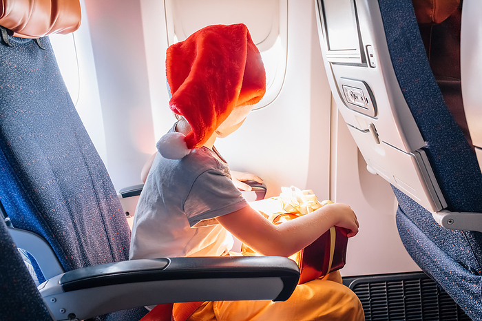 Child in Santa Claus hat flies at cabin of airplane with holiday gift., by Cavan Images / Kseniia Liubasova