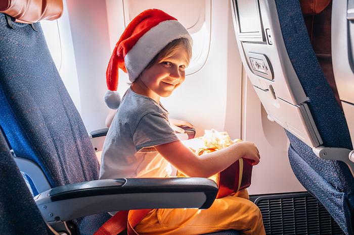 Child in Santa Claus hat flies at cabin of airplane with holiday gift., by Cavan Images / Kseniia Liubasova
