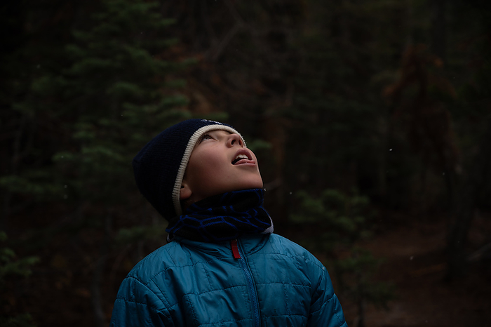 Boy in winter hat and coat sticking out tongue to catch snowflakes, by Cavan Images / Liz Celeste