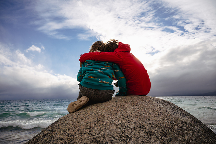Brothers sitting on boulder hugging and looking out at lake, by Cavan Images / Liz Celeste