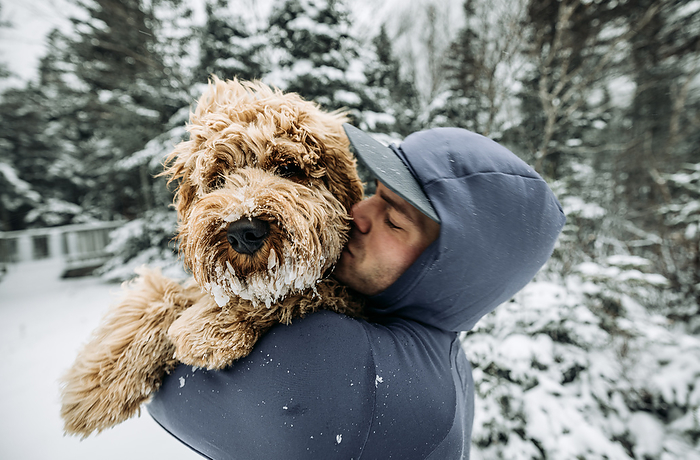 Man kisses his snow covered dog while walking in snow covered woods, by Cavan Images / Chris Bennett