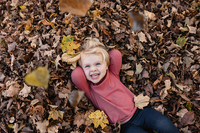 Smiling child laying in leaf pile as autumn leaves fall, by Cavan Images / Krista Taylor