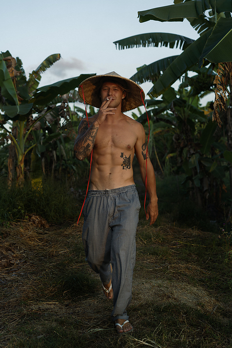 A young man wearing an Asian cone hat in rice fields at sunset. Bali., by Cavan Images / Yuliya Kirayonak