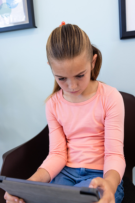 Focused caucasian girl patient using tablet sitting in hospital waiting room. Communication, childhood, lifestyle, medical services, healthcare and hospital, unaltered.