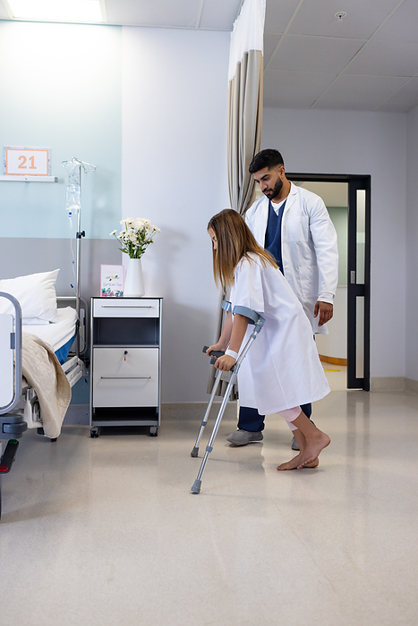 Diverse male doctor observing girl patient use crutches in hospital ward, copy space. Injury, mobility, childhood, medical services, healthcare and hospital, unaltered.
