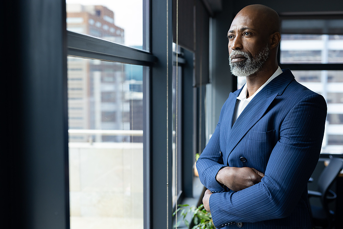 Thoughtful senior african american businessman standing looking out of window in office, copy space. Work, contemplation, business and office, unaltered.