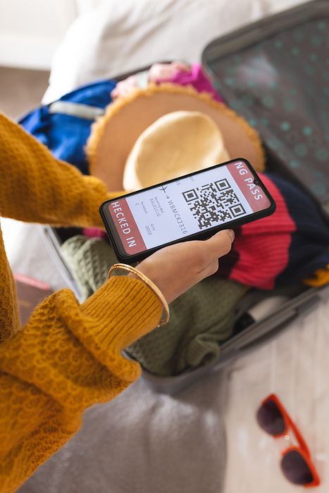 Hands of biracial woman using smartphone and packing suitcase at home. Lifestyle, travel, vacation, communication and domestic life, unaltered.