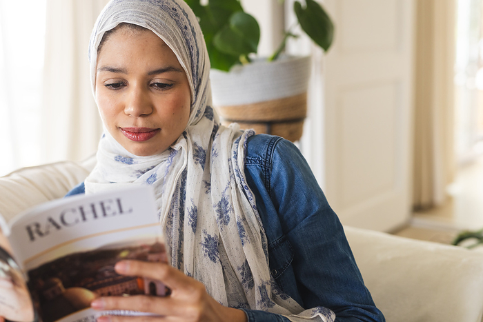 Biracial woman in hijab reading book in living room. Lifestyle, free time and domestic life, unaltered.