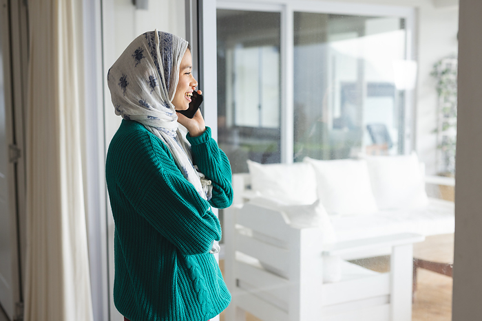 Happy biracial woman in hijab talking on smartphone at sunny home. Lifestyle, communication and domestic life, unaltered.
